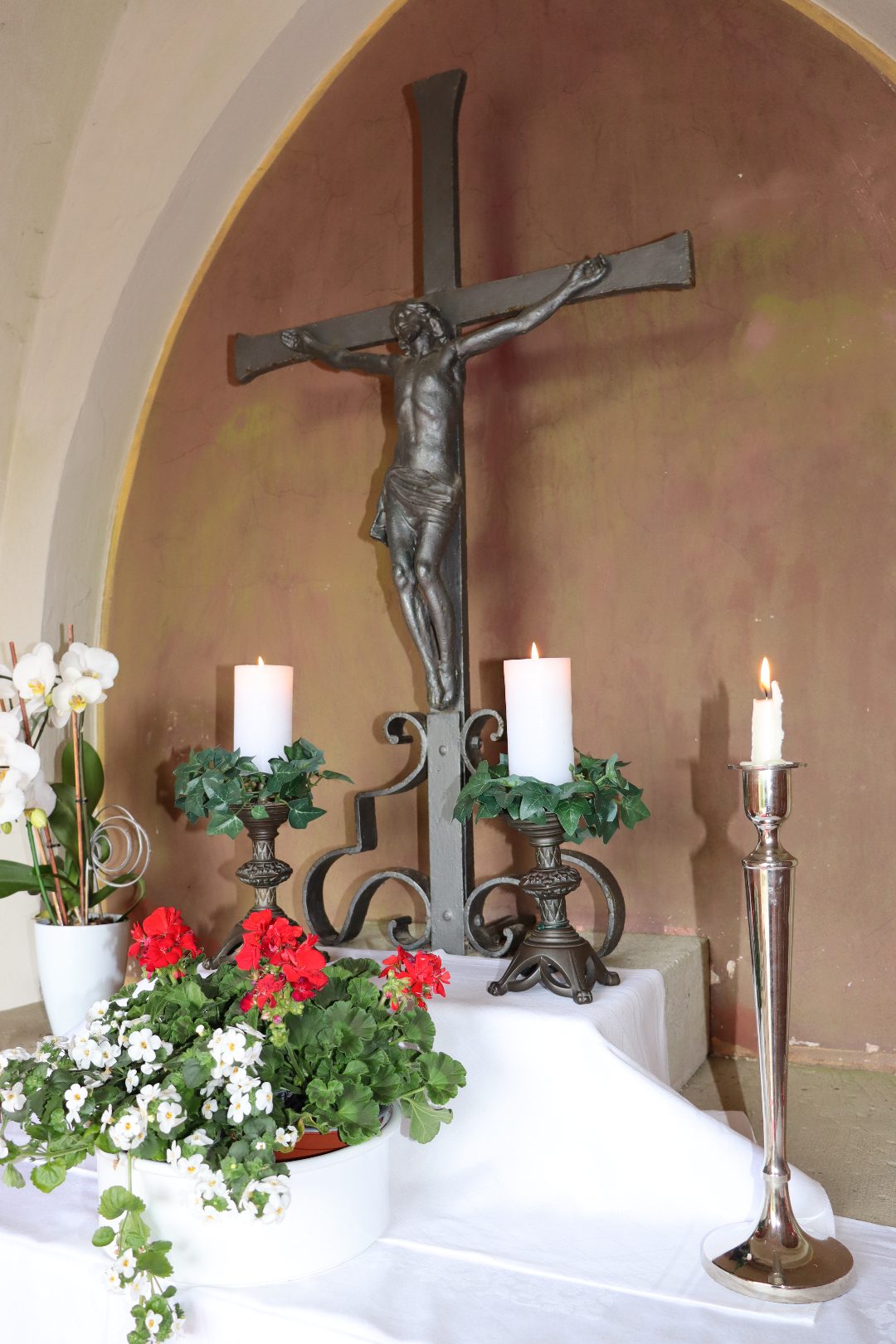 Detailansicht Altar: eisernes Kreuz mit Christuskorpus, Kerzenleuchter und roten Geranien