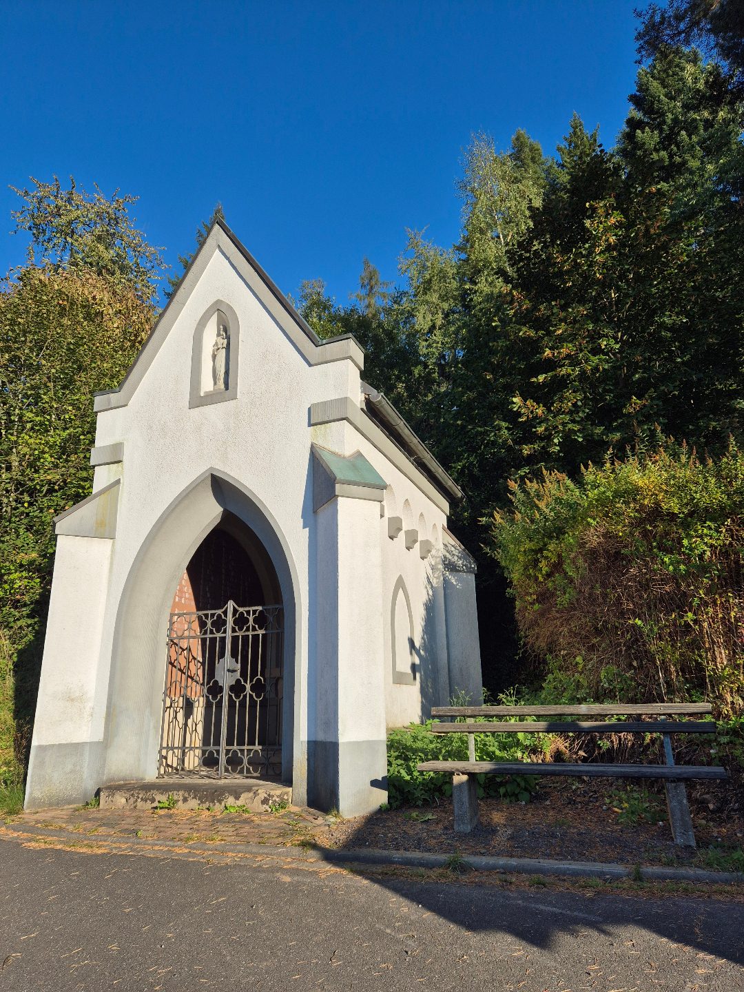 Weiße Pestkapelle mit neogotischer Gitterpforte und Holzbank, Herbst 2025